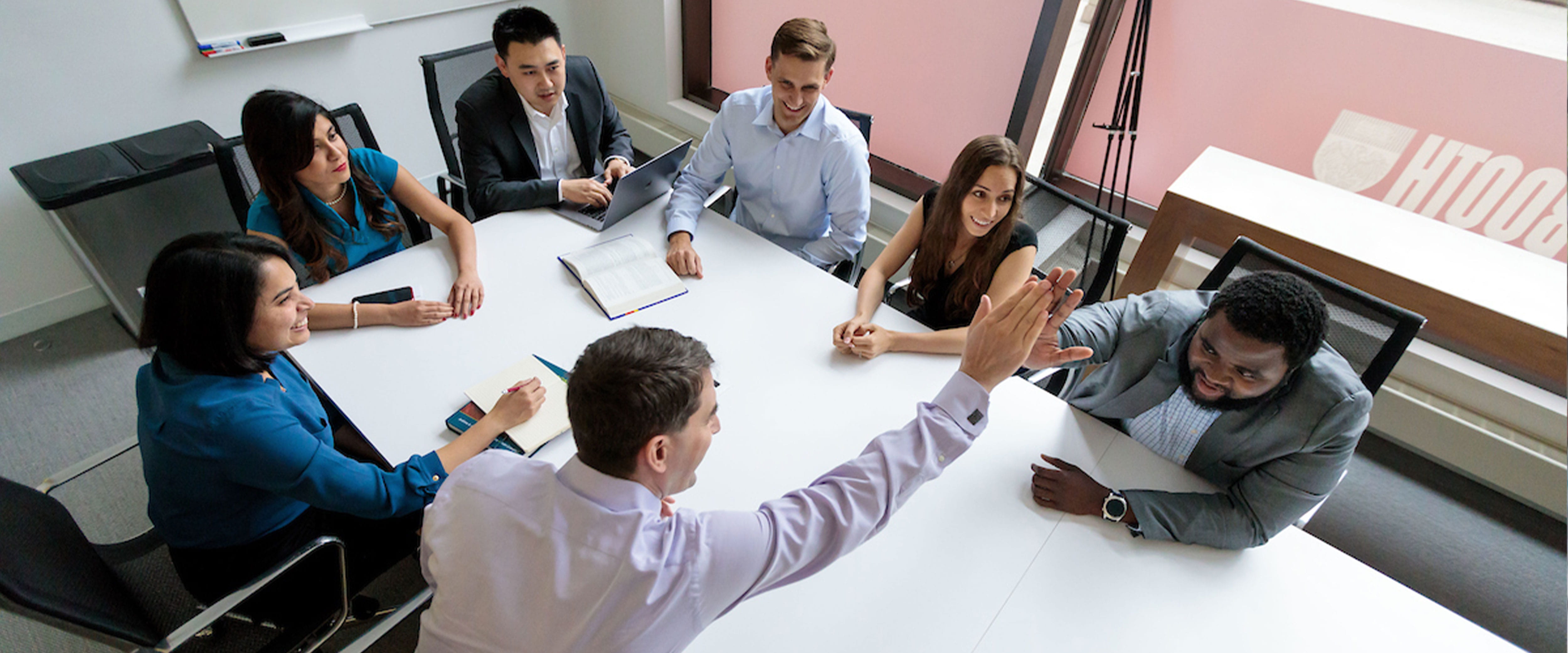 Group of students meeting at a conference table, two students high fiving