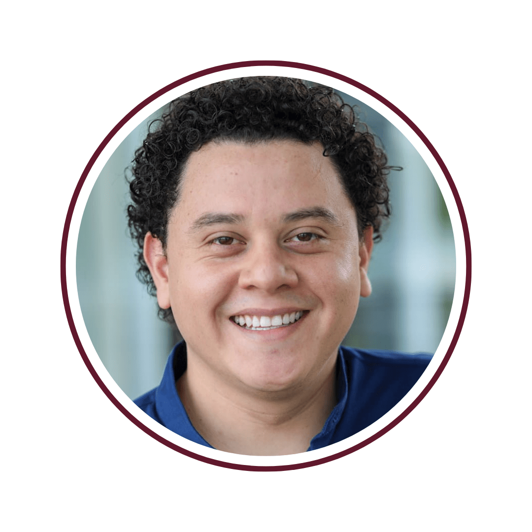 Young man smiling in front of atrium windows. He has dark curly hair and is wearing a blue shirt. 